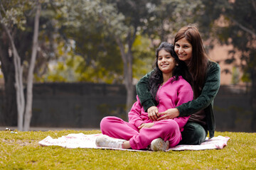 Indian mother and teenage girl sitting together in garden , Mother hug daughter while sitting in park