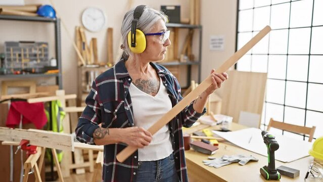Serious, grey-haired middle age woman carpenter deeply invested in her woodwork, fervently looking at a timber plank in her homely carpentry workshop