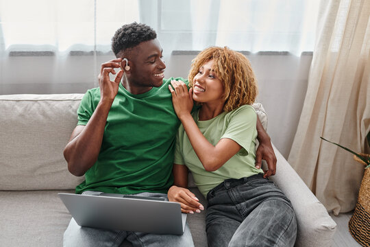 happy african american man in hearing aid device sitting on couch with girlfriend and using laptop