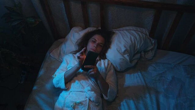 Insomniac young woman lying in bed scrolling smartphone at night. Top view shot.