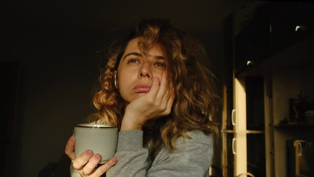 Creative crisis. Morning portrait of young woman. Lady holds mug of morning coffee, sighs heavily while sitting in sunny room at sunrise.