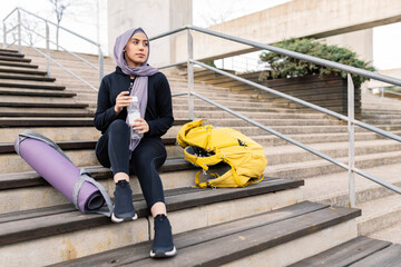 Sporty woman with headscarf sitting on the stairs after exercising contemplating the horizon with...