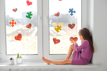A lonely girl is sitting on the windowsill by a window decorated with puzzles for Autism Awareness...