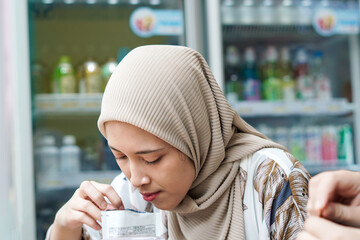 Indonesian Hijab Woman Pouring Packaged Beverage into Cup with Ice Cubes at Cafe or Minimarket - obrazy, fototapety, plakaty