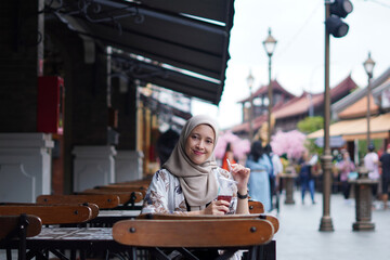 Obraz premium Indonesian Hijabi Woman Smiling with Refreshment at Cafe Bench. Joyful Moments at the cafe concept.