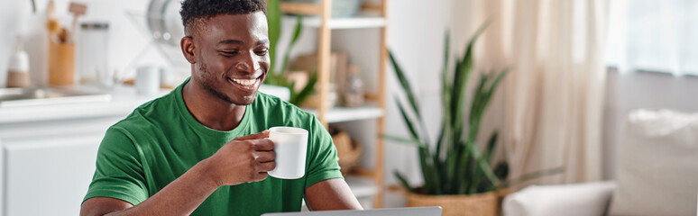 Smiling african american man enjoying cup of coffee and smiling in kitchen, horizontal banner