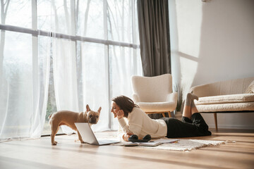 In sunlight. Lying down on floor with laptop. Looking at pet. Young woman is with her pug dog at home - obrazy, fototapety, plakaty