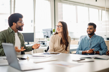 A spirited discussion unfolds in a light-filled office, where a man in a green shirt gestures animatedly, embodying a dynamic exchange of ideas among a diverse professional team