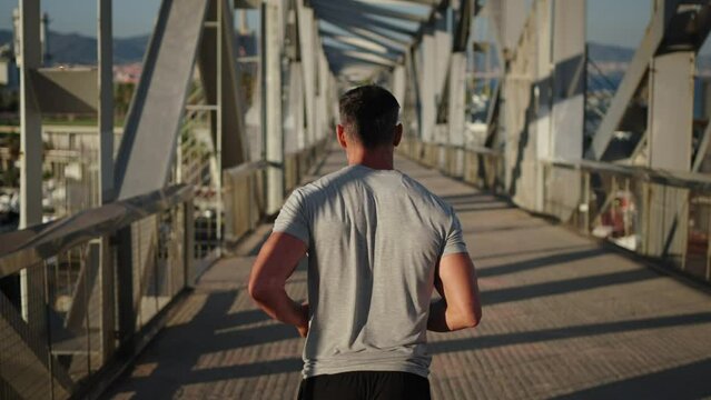 Follow back of unrecognizable mature strong man running alone on modern urban bridge, disciplined male stays fit and firm, burning calories for healthy body. People doing daily cardio sports routine