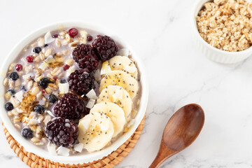 Oatmeal bowl with banana, berries, granola, coconut flakes, and chia seeds with wooden spoon isolated on white marble table. Close-up. Healthy vegan plant-based breakfast meal.
