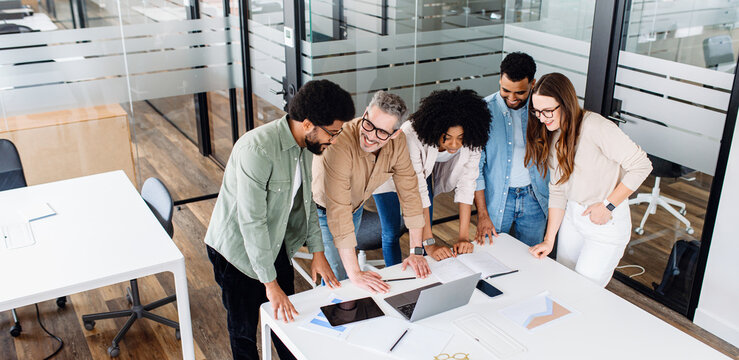 A diverse team is huddled around a table in the office, reviewing documents, creating a vision of the new project, and discussing strategies, reflecting a dynamic brainstorming session.