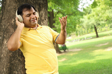 Portrait of obese Indian man lost in music in calm green city park using headphones early morning...