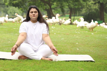 A obese young lady exercising, doing Yoga poses in green serene, peaceful environment early morning in park maintaining healthy lifestyle. Concentration. Focus. International Yoga day. Meditating.