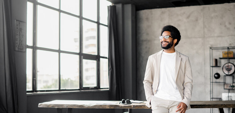 A young Brazilian businessman casually leans at desk in a spacious office, his bright smile exuding confidence and ease in the modern workspace. The panoramic windows fill the room with natural light - Powered by Adobe