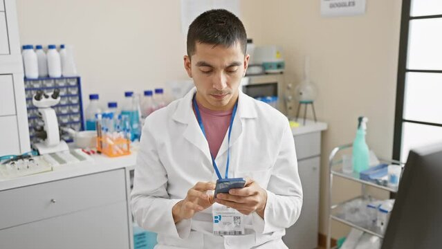 Confident young hispanic man, a scientist in lab uniform, showing a serious no-go hand gesture, holding smartphone, symbolising strong defense at laboratory.