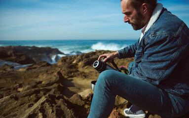 Caucasian handsome relaxed bearded man tourist pouring hot tea or coffee from thermos into a mug, resting by ocean