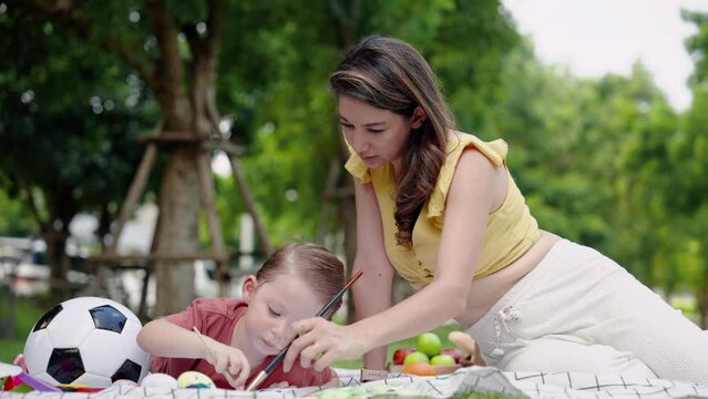 Beautiful Caucasian woman, young mother, sits near handsome son lying on small mat, mother son do activities together by having picnic in garden, sitting coloring with their son on nice day.