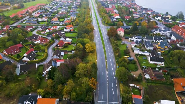 Herbstliche Vogelperspektive auf eine lange, gerade Stra&szlig;e durch eine Vorstadt mit Blick auf den See. Drohnenaufnahme. Landstra&szlig;e, Egernsundbr&uuml;cke, Egernsund, D&auml;nemark.