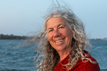 Beaming grey-haired older woman posing on a beach, looking at camera with delighted, happy smile,...