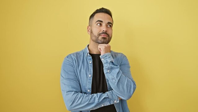 Young hispanic man wearing denim shirt standing with hand on chin thinking about question, pensive expression. smiling and thoughtful face. doubt concept. over isolated yellow background