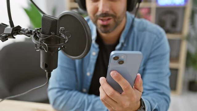 Hispanic man with beard in studio using smartphone and microphone for podcast recording.