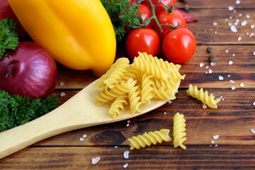 On a wooden table there is pasta in a wooden spoon against a background of vegetables.
