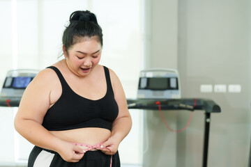 Plus-size Asian woman exercise in gym, Joyful attire proudly measures her waist, showcasing positive results with a treadmill backdrop. fitness gear smiling while measuring waistline