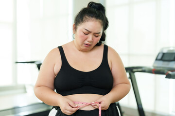 Plus-size Asian woman exercise in gym, visibly upset while measuring waist, moment of struggle on fitness journey, Emotionally overwhelmed with measuring tape, confronting weight loss challenges