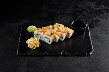 Shrimp roll with shrimp on top and cucumber and cheese inside, on a black slate with soy sauce, wasabi, and ginger