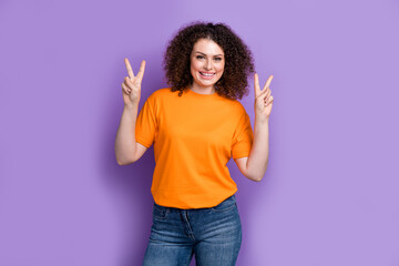 Photo of cute positive woman wear orange t-shirt smiling showing two v-signs isolated purple color background