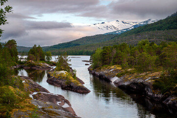 In the evening, the setting sun illuminates the snowy peaks of the mountains. A lake with a rocky shore in the countryside in Norway.