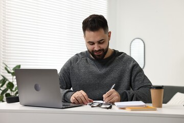 E-learning. Young man taking notes during online lesson at white table indoors