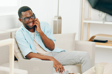 Smiling African American Man Working on Laptop in Modern Apartment, Making Phone Call for Online...