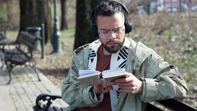 Young handsome hipster freelance business man wearing jacket reading a book while sitting outdoors. Businessperson on lunch break listening music on wireless headset and read novel to clear his mind