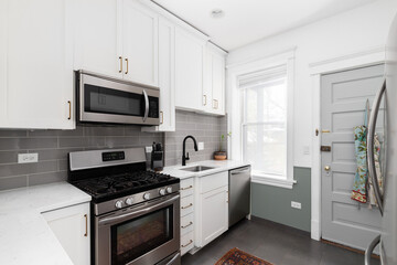 A small, white kitchen with a grey subway tile backsplash, stainless steel appliances, and grey...