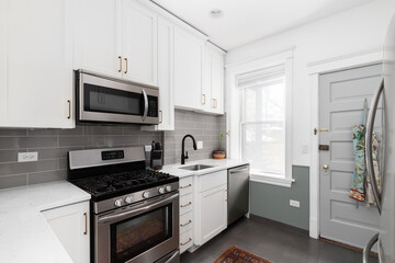 A small, white kitchen with a grey subway tile backsplash, stainless steel appliances, and grey tiled floor. No brands or labels.