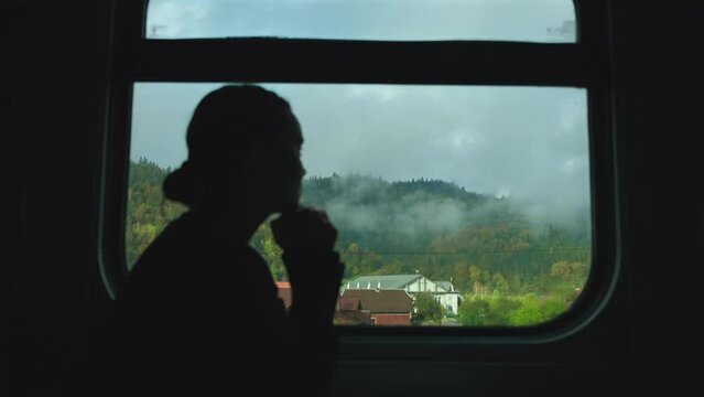 Woman silhouette near the window in the train compartment. View on the mountains and small village through train window, fog over mountain peaks in the morning