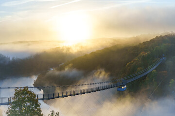 Blick zur Hängebrücke Titan RT an der Rappbodetalsperre im Harz. Die Sonne scheint über die Wipfel und taucht die nebelverhangene Landschaft über dem Bodetal in ein goldenes Licht. - obrazy, fototapety, plakaty