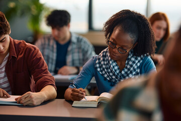 Black college student writing in her notebook while studying in classroom.