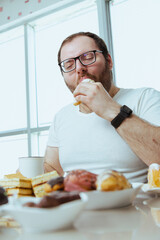 Man sitting in front of a table full of sweet food, eating pastry with cream. Fat man sugar addict, can't say no and stop eating, weight loss problem. Depressed man eats a lot of unhealthy meal.