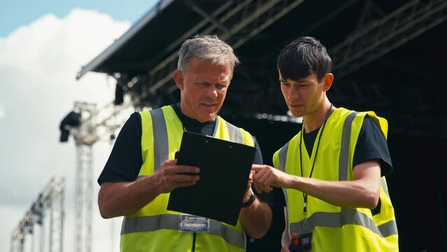 Male Production Team With Clipboard Setting Up Outdoor Stage For Music Festival 