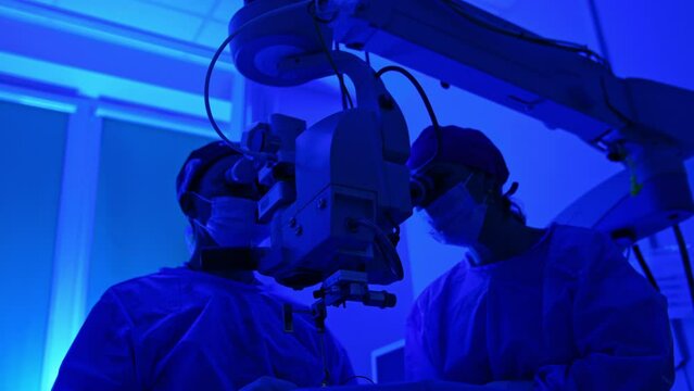 Two medics stand looking at the microscope in operational room with blue light. Low angle view on the surgeons performing eye surgery.