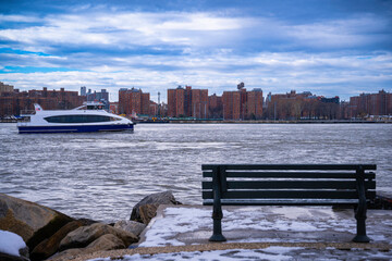 Lower Manhattan Skyline with Red Brick Buildings, Cruising Ferry, an empty wooden bench, and snow on the jetty along the East River in Brooklyn, New York, USA