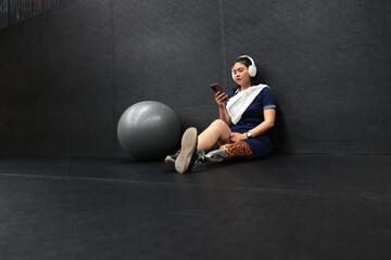 A confident woman with a leg prosthesis and wearing headphones sits on the floor listening to her smartphone's music. after exercising at the gym