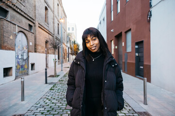 Fototapeta na wymiar Portrait of a cheeful young black woman with a coat walking on a narrow street.