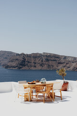 A serene outdoor dining setup in Santorini overlooks the stunning caldera. The wooden table and chairs contrast with the whitewashed terrace, creating a perfect Mediterranean scene.