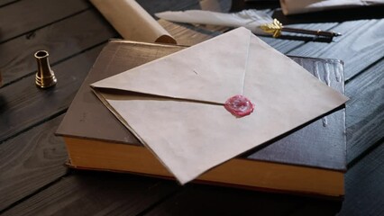 Shot of the desk with old paper envelope. Vintage love letter laying on the book sealed with wax stamp, feather quill and lighted candle around.