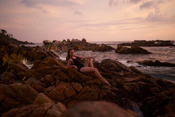 Woman relaxes on coastal boulders, serene dusk light bathing scene. She gazes at ocean horizon, waves crash gently. Twilight peace envelops, reflecting travel, wellbeing, natures splendor.