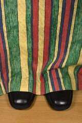 Black dress shoes peeking from behind colorful curtains on beige hard wood floor, large detailed...
