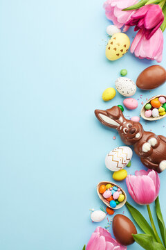 Easter joy: a bounty of sweets. Top view vertical shot of tulips and a delightful assortment of Easter chocolates and candies on a sky blue surface with space for heartfelt Easter wishes